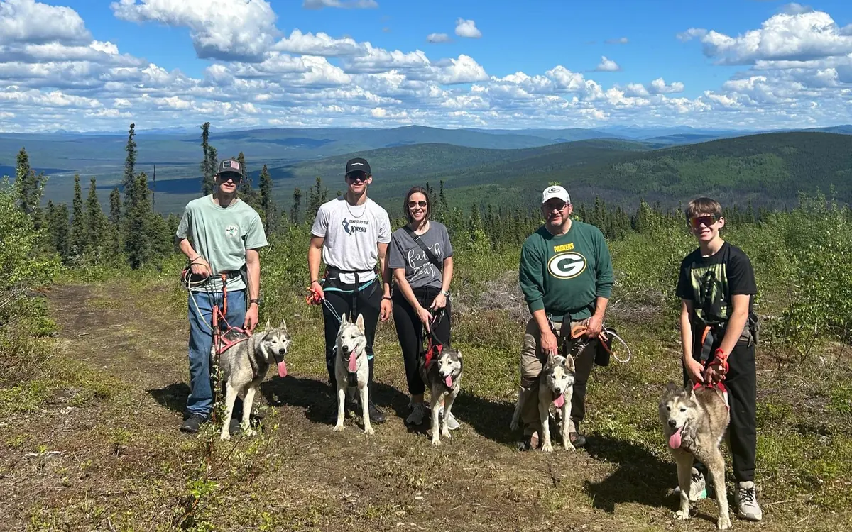 Five visitors on a summer hilltop near Fairbanks, each holding a Siberian husky on a line, with forested hills stretching to the horizon under a blue sky