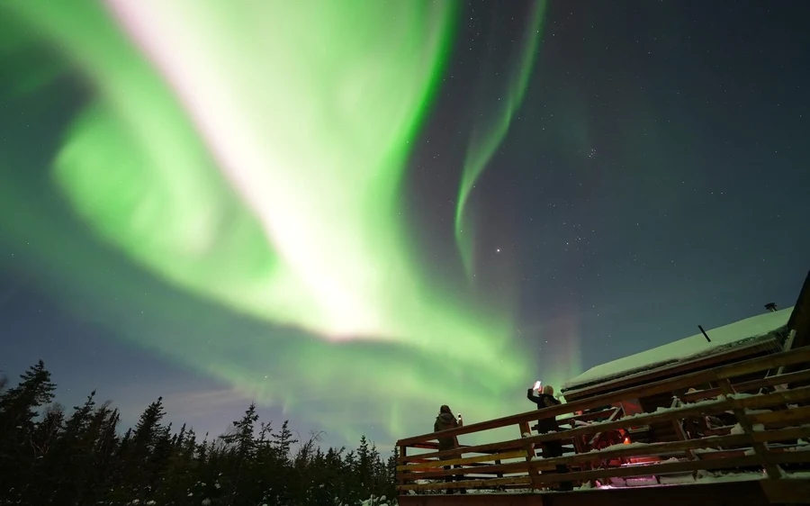 A green aurora over a cabin deck with people watching from the rail